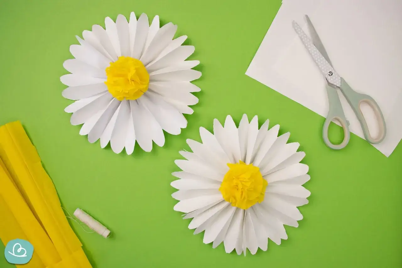 Zwei weiße Papierblumen mit gelben Zentren, bereit zum Basteln. Schere und gelbes Krepppapier liegen daneben.