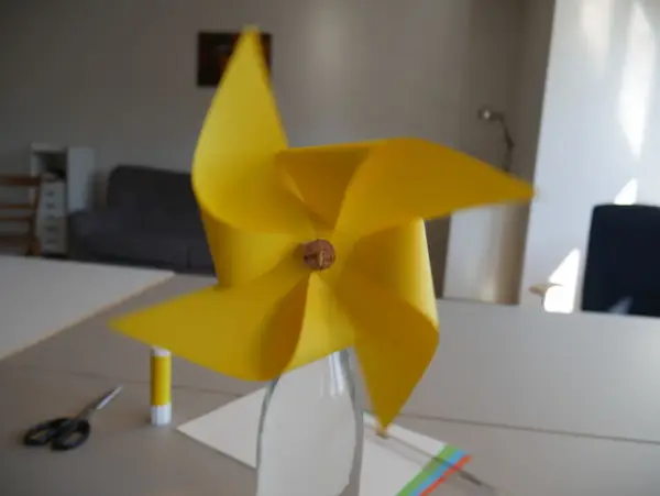 Ein gelbes Windrad, das man leicht selber bauen kann, steht in einer Glasflasche. Schere und Klebestift liegen bereit.